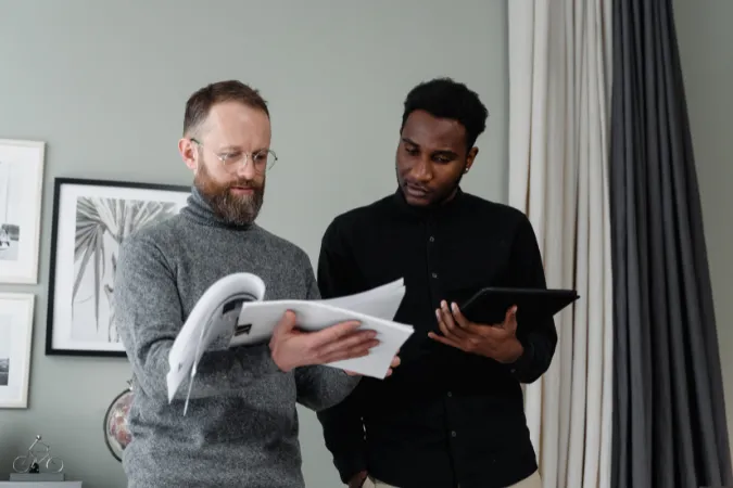 Dois homens analisando relatórios impressos e dados em tablet durante uma reunião.