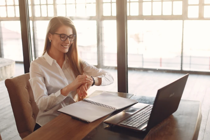 Profissional sorrindo enquanto revisa o planejamento do dia com um caderno e notebook sobre a mesa, representando equilíbrio entre foco e leveza.