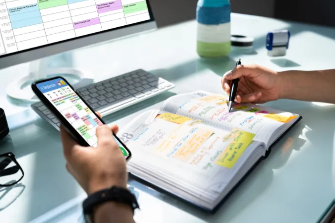 Pessoa organizando cronograma de projetos com planner físico, smartphone e calendário digital.