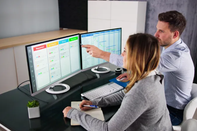 Dois profissionais organizando o cronograma de projetos com quadros Kanban em monitores.