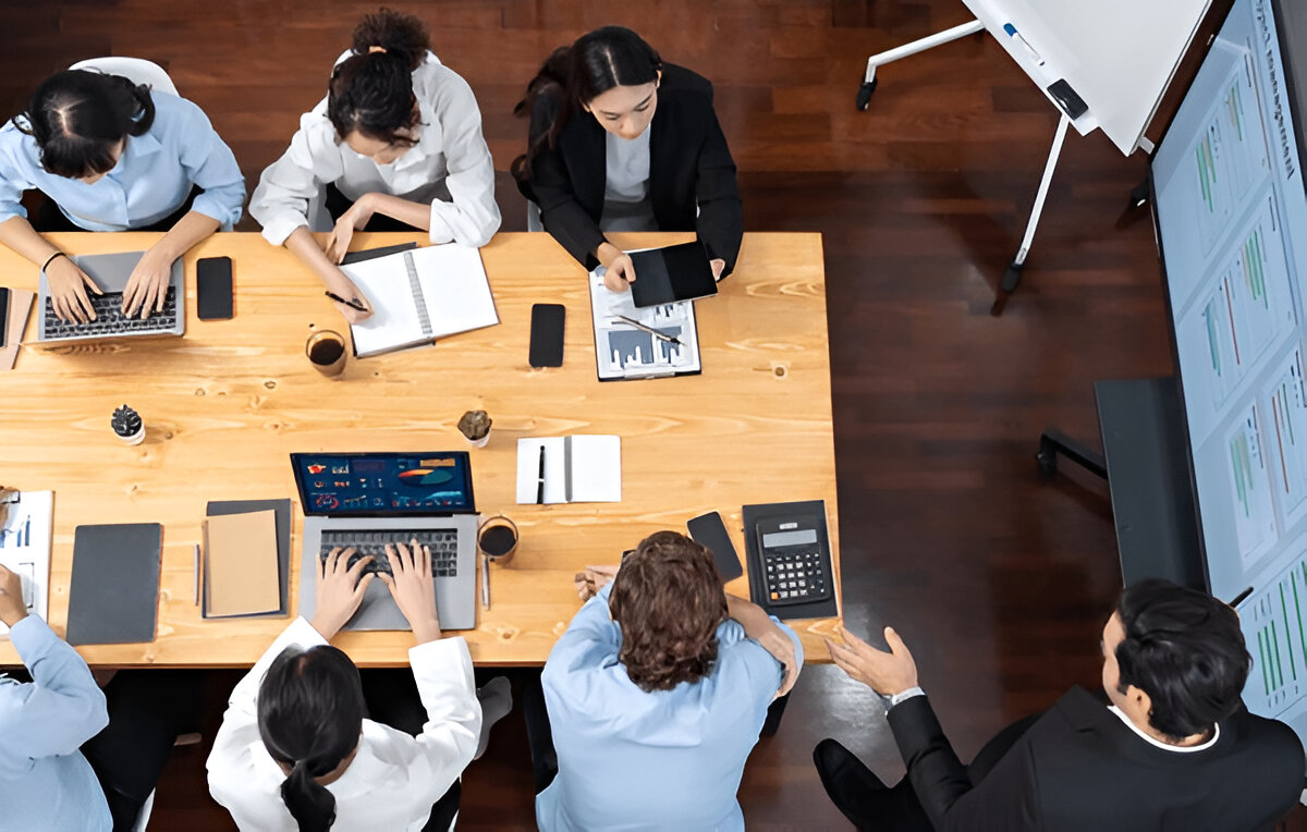 Equipe reunida em mesa de trabalho analisando dados e cronogramas apoiados por gráfico de Gantt projetado na tela.