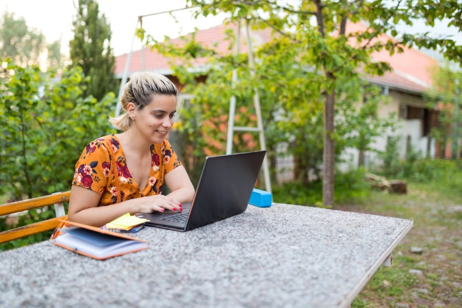 Mulher trabalhando em um laptop ao ar livre, sentada em uma mesa no quintal, representando flexibilidade no home office.