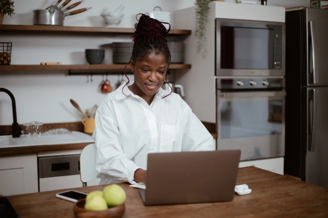 Mulher usando laptop sobre a mesa da cozinha enquanto trabalha de casa, ilustrando adaptação de espaços domésticos para o trabalho remoto.