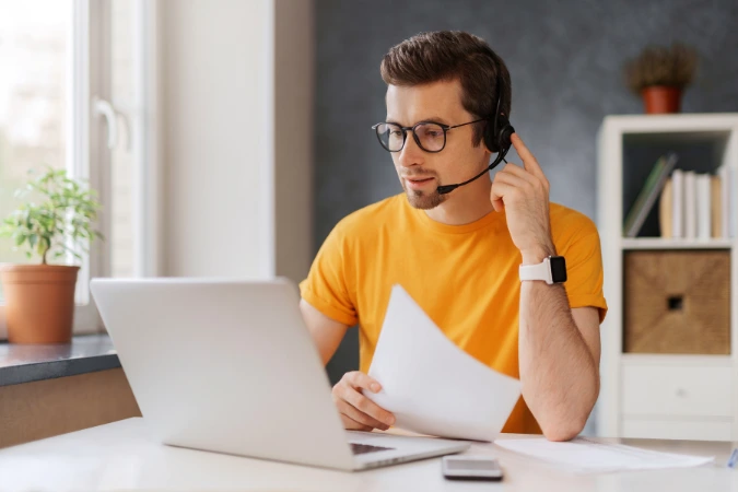Homem trabalhando no laptop com headset, participando de uma chamada de vídeo e segurando documentos.
