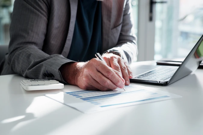 Empresário analisando planilhas financeiras e preenchendo dados de fluxo de caixa ao lado de notebook.