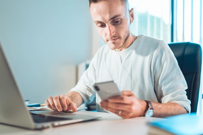Profissional trabalhando remotamente enquanto gerencia tarefas pelo celular e computador