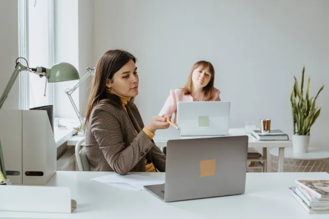 Duas profissionais analisando atividades em laptops, representando alinhamento e colaboração com apoio de um cronograma de projeto.