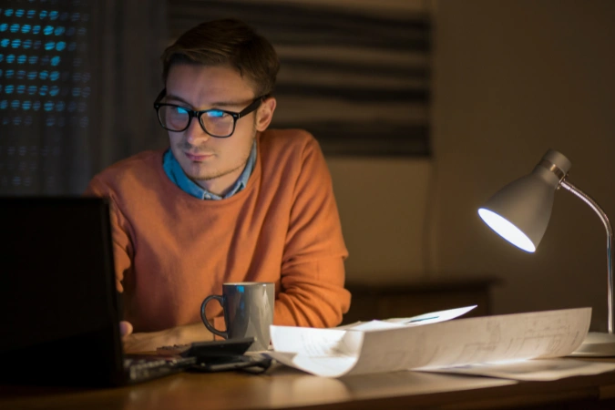 Homem concentrado no computador à noite com papéis e xícara de café, ilustrando a importância da gestão de tempo.
