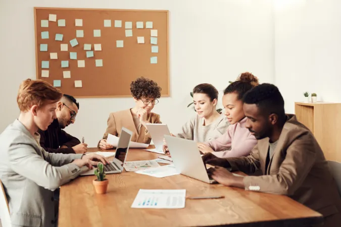 Seis profissionais em mesa de reunião com laptops, papéis e post-its organizando planos de trabalho.