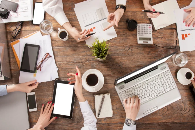 Mesa com notebooks, tablets e papéis de relatórios empresariais integrando dados de diferentes setores.