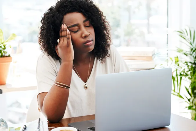Mulher tocando a cabeça em frente ao computador, simbolizando dores e estresse relacionados à sobrecarga de trabalho.