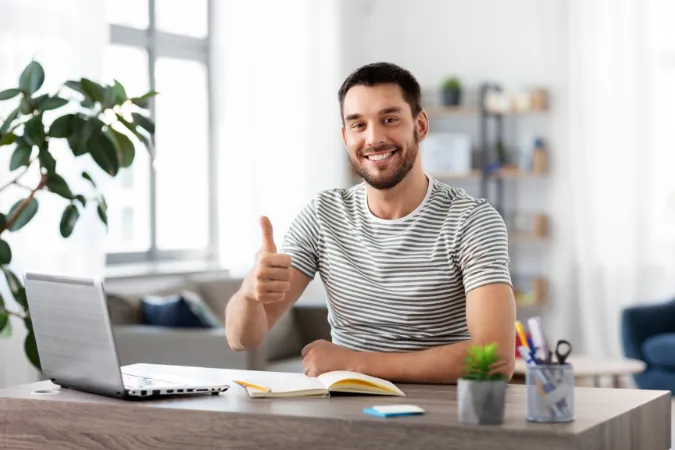 Homem sorridente fazendo sinal de positivo em frente ao notebook, representando motivação e produtividade ao evitar a sobrecarga de trabalho.