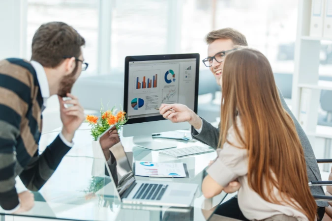 Equipe reunida discutindo gráficos de desempenho apresentados em um monitor para identificar e corrigir lacunas estratégicas.