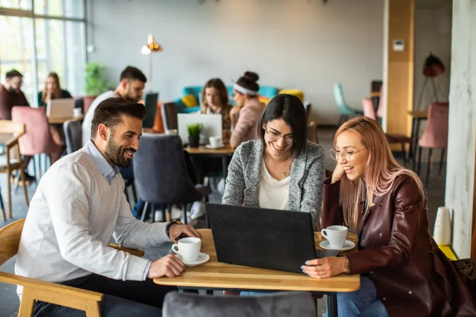 Três colegas em um café, reunidos em torno de um notebook, revisando tarefas e prioridades com foco em Autogestão no trabalho.
