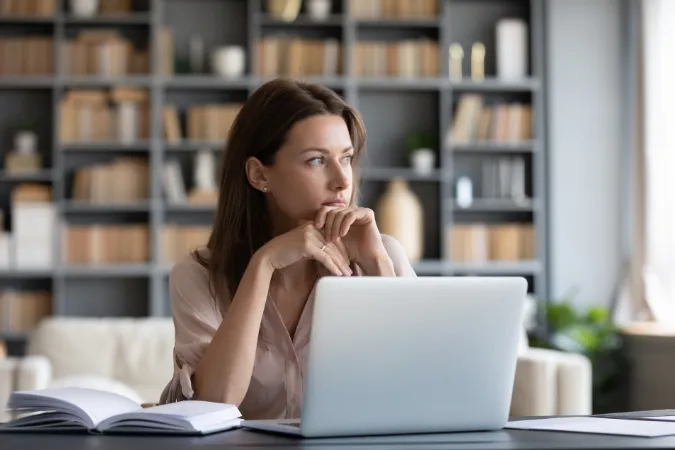 Mulher em frente ao laptop, em um momento de ociosidade no trabalho, pensando em como retomar o fluxo de atividades.