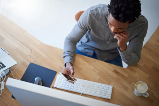 Homem em frente ao computador e ao celular, em período de ociosidade, buscando organizar tarefas para retomar a produtividade.