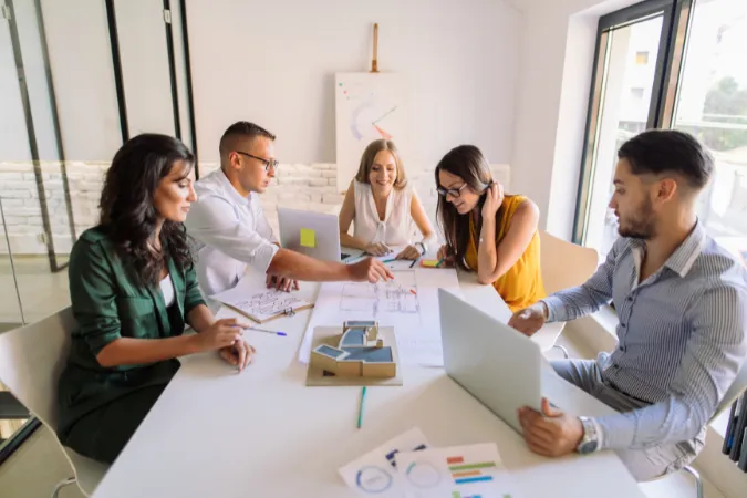Equipe reunida ao redor de uma mesa discutindo projetos com papéis, laptops e maquetes