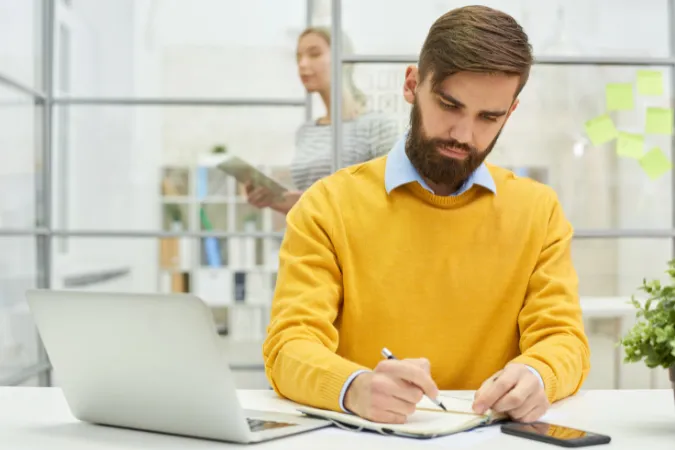 Homem escrevendo em caderno com laptop e celular sobre a mesa, em ambiente corporativo