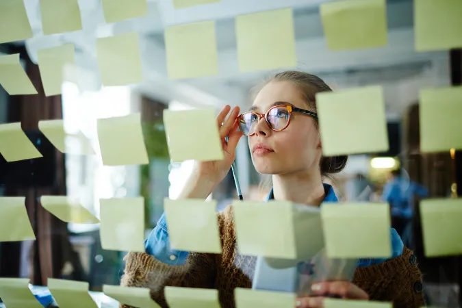 Profissional analisando diversos post-its em um painel de vidro, refletindo sobre prioridades e tarefas no planejamento de projetos.