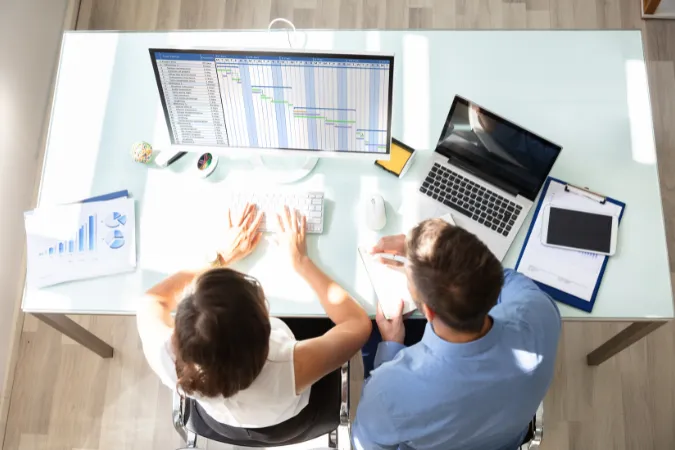 Dois profissionais trabalhando em frente a um computador com cronograma de projeto em formato de gráfico de Gantt.