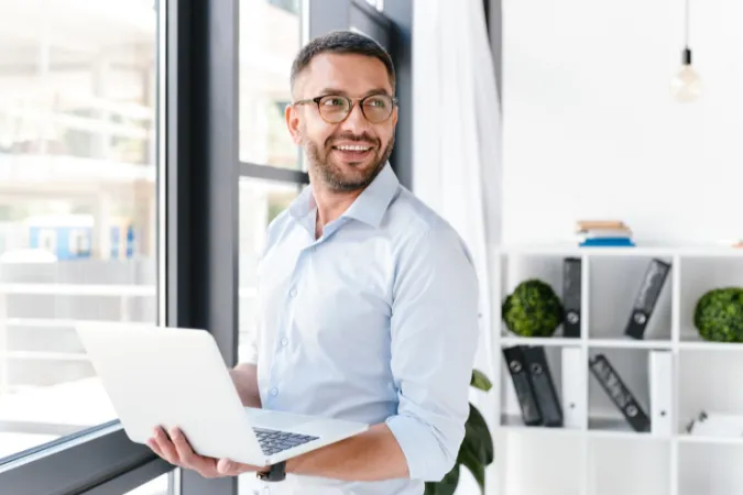 Profissional sorridente em escritório utilizando notebook para acessar software de gestão.