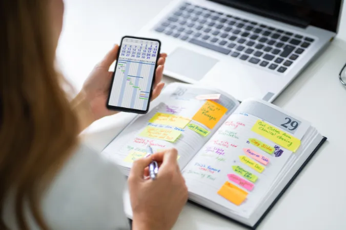 Pessoa analisando um gráfico de Gantt no celular e uma agenda física para alinhar o gerenciamento do cronograma do projeto.