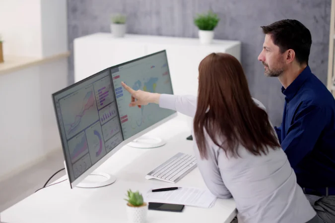 Equipe analisando dados em tela dupla de computador, discutindo indicadores e resultados de projetos