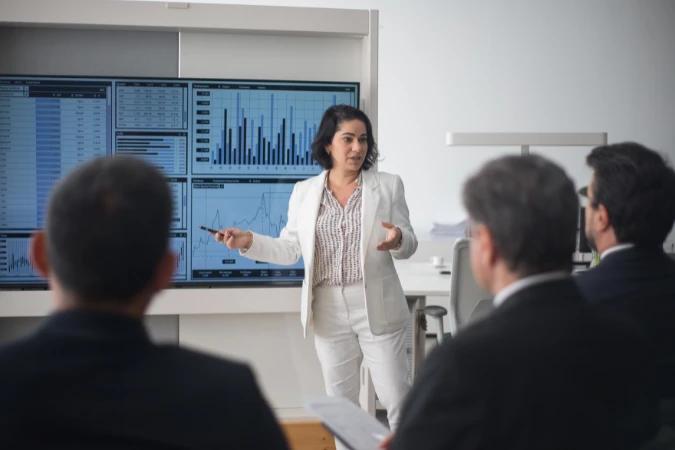 Gestora apresentando gráficos e relatórios em telas digitais para executivos em reunião.