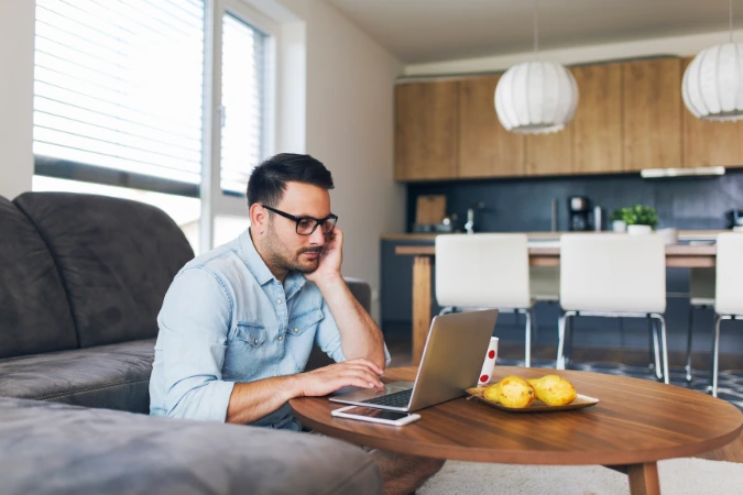 Homem trabalhando de casa usando laptop e smartphone, representando a rotina do trabalho remoto com apoio de ferramentas digitais.