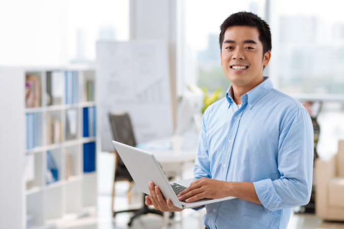Homem sorridente segurando notebook em escritório moderno, representando segurança financeira ao adotar um software de gestão.