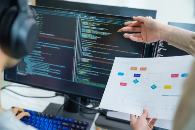 Duas pessoas analisando fluxograma impresso enquanto observam código no monitor para identificar gargalos no desenvolvimento de software.
