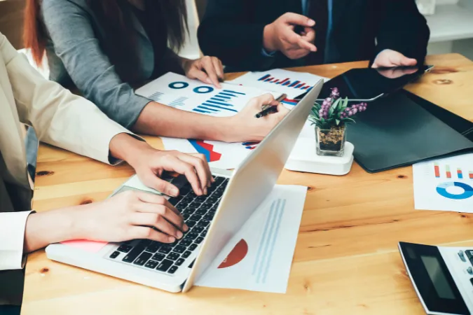 Profissionais analisando relatórios e gráficos em laptops e documentos sobre a mesa em reunião de projetos.