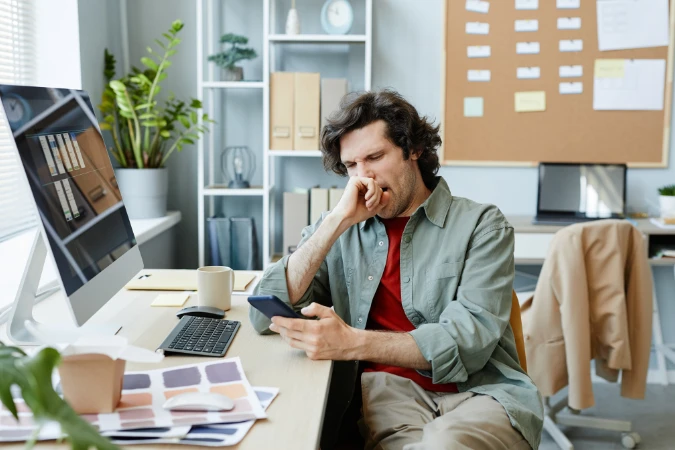 Homem em frente ao computador, bocejando e distraído com o celular, retratando a queda de foco em ambientes que estimulam a procrastinação.