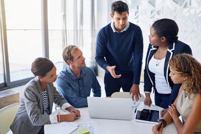 Equipe diversa reunida em mesa de trabalho, discutindo estratégias com notebook e tablet, representando colaboração entre áreas em projetos