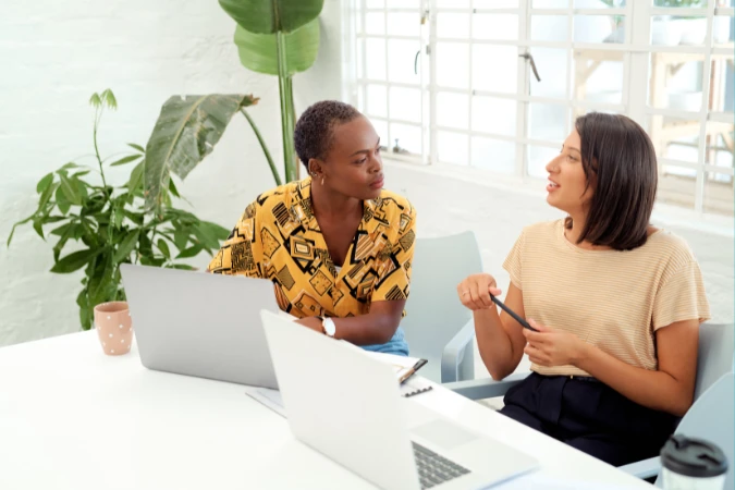 Duas profissionais conversando e trocando ideias diante de notebooks, simbolizando feedback e adaptação na gestão antifrágil.