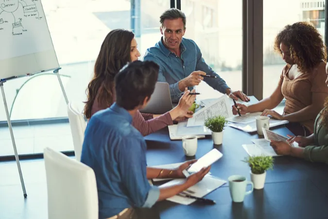 Mãos de profissionais apontando para tela com cronograma enquanto discutem a distribuição de tarefas em equipe multidisciplinar.