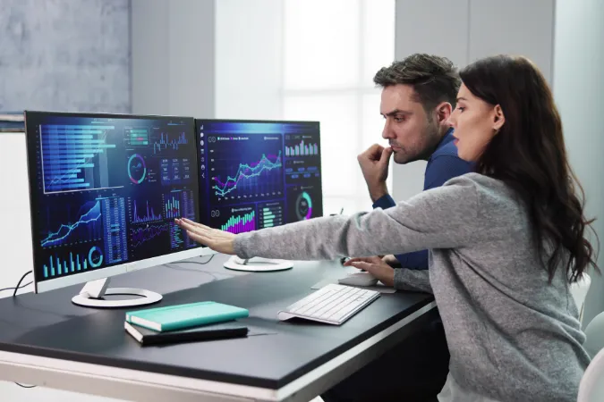 Dupla de profissionais analisa gráficos e métricas em um dashboard de projetos exibido em dois monitores.