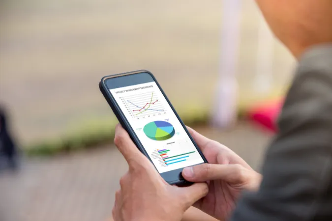Profissional visualiza gráficos e relatórios de desempenho de projetos em dashboard acessado por smartphone.