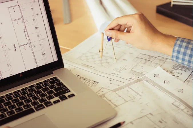 Mão com compasso sobre planta técnica ao lado de notebook com projeto digital