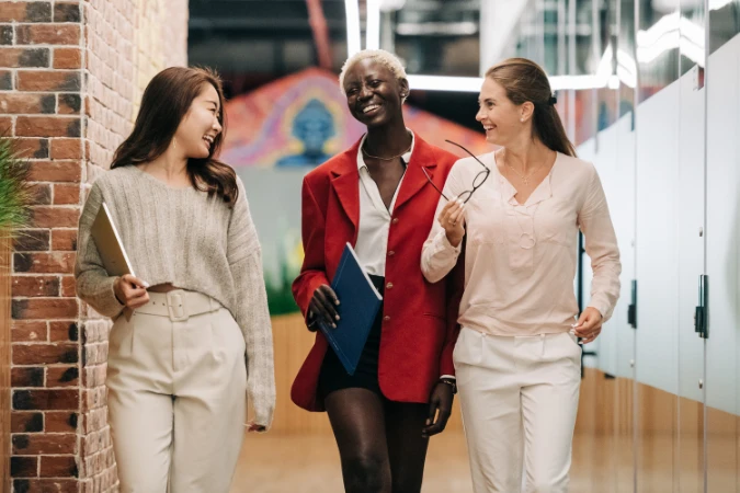 Três mulheres caminhando pelo escritório sorrindo e conversando de forma descontraída.