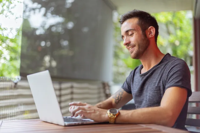 Profissional sorridente utilizando notebook em ambiente externo para analisar informações de desk research organizadas em software de gestão.