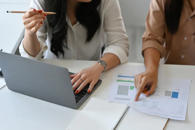 Duas profissionais analisando relatórios financeiros em laptop e planilhas impressas, representando o uso de software de gestão para rateio de custos.