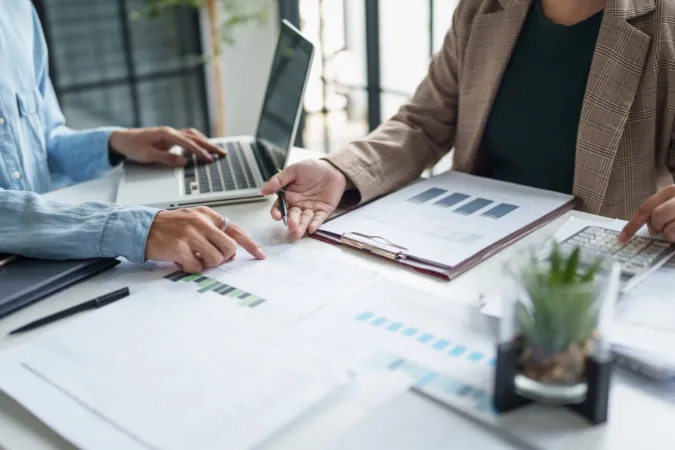 Duas pessoas trabalhando lado a lado, revisando gráficos financeiros e indicadores de projeto sobre a mesa.