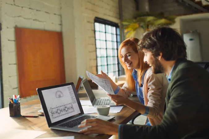 Dois arquitetos analisando plantas no notebook e discutindo ajustes no projeto durante uma reunião descontraída.