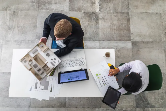 Visão aérea de dois profissionais avaliando uma maquete arquitetônica e plantas digitais em uma mesa de trabalho.