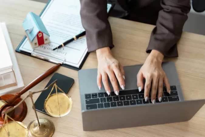 Profissional digitando em um laptop com documentos e planilhas ao redor, representando análise de horas e desempenho.