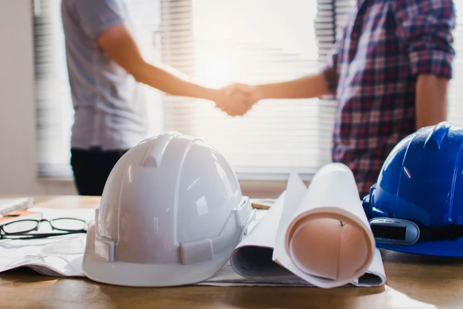 Documentos e capacetes sobre a mesa com pessoas apertando as mãos, simbolizando aprovação e validação do projeto arquitetônico.