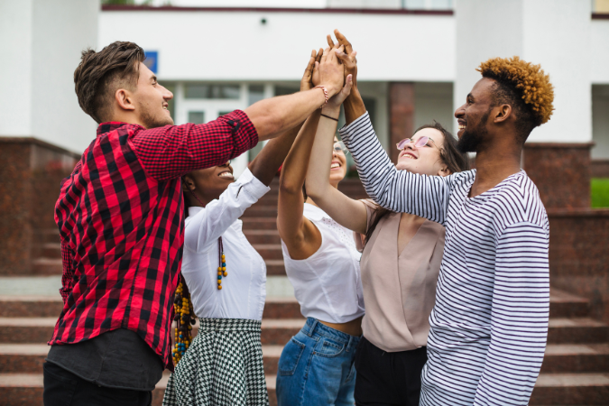 Grupo diverso de jovens sorridentes realizando um gesto coletivo de "high five" ao ar livre, representando espírito de equipe, celebração e união.