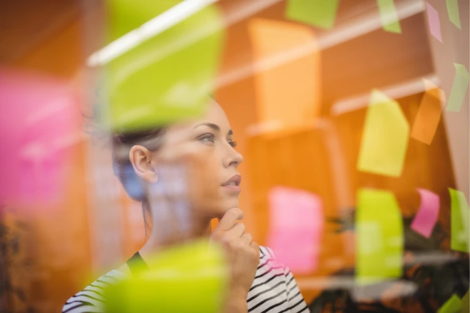 Mulher pensativa observa post-its coloridos fixados em uma parede de vidro, em ambiente iluminado, simbolizando uma etapa de análise e ideação no processo de Design Thinking.