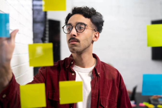 Homem jovem, de camisa vermelha e óculos, analisa post-its coloridos colados em um painel de vidro, representando um momento de brainstorming e ideação característico do Design Thinking.